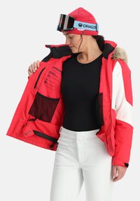 Rode en witte skijas met bontafwerking, ritsen en meerdere binnenzakken. Draagt open, gecombineerd met een zwart topje en witte broek.