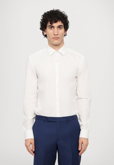 Chemise blanche à manches longues avec un col classique, fermeture à boutons et une texture subtile, associée à un pantalon cintré bleu marine.