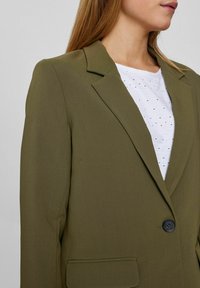 Õlivihre ülikond, millel on struktureeritud õlad, üks must nööbiklapp ja kaks eesmist taskut. Kanna valge päeva kleidi peal, millel on väikesed detailid.