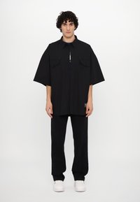 Black oversized button-up shirt with two chest pockets, paired with black pants and white sneakers. Smooth texture, straight cut.