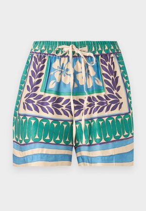 Naine seisab, kandes mustriga lühikeste varrukatega särki ja sobivaid lühikesi pükse beežide sandaalidega ühtlasel taustal.