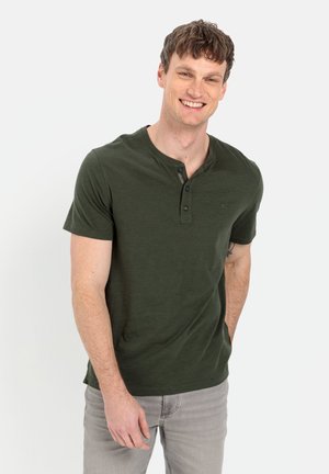 Chemise henley verte à manches courtes avec un plastron à trois boutons et un col rond, associée à un jean gris clair. Coupe décontractée et texture douce.