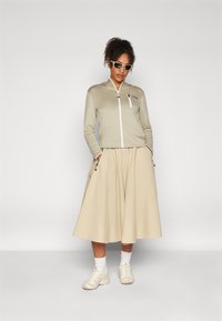 Beige cipzáros dzseki fehér díszítéssel, bézs midi szoknya és krémszínű tornacipő. Hullámos haj, világos napszemüvegben. Sima szövettextúra.