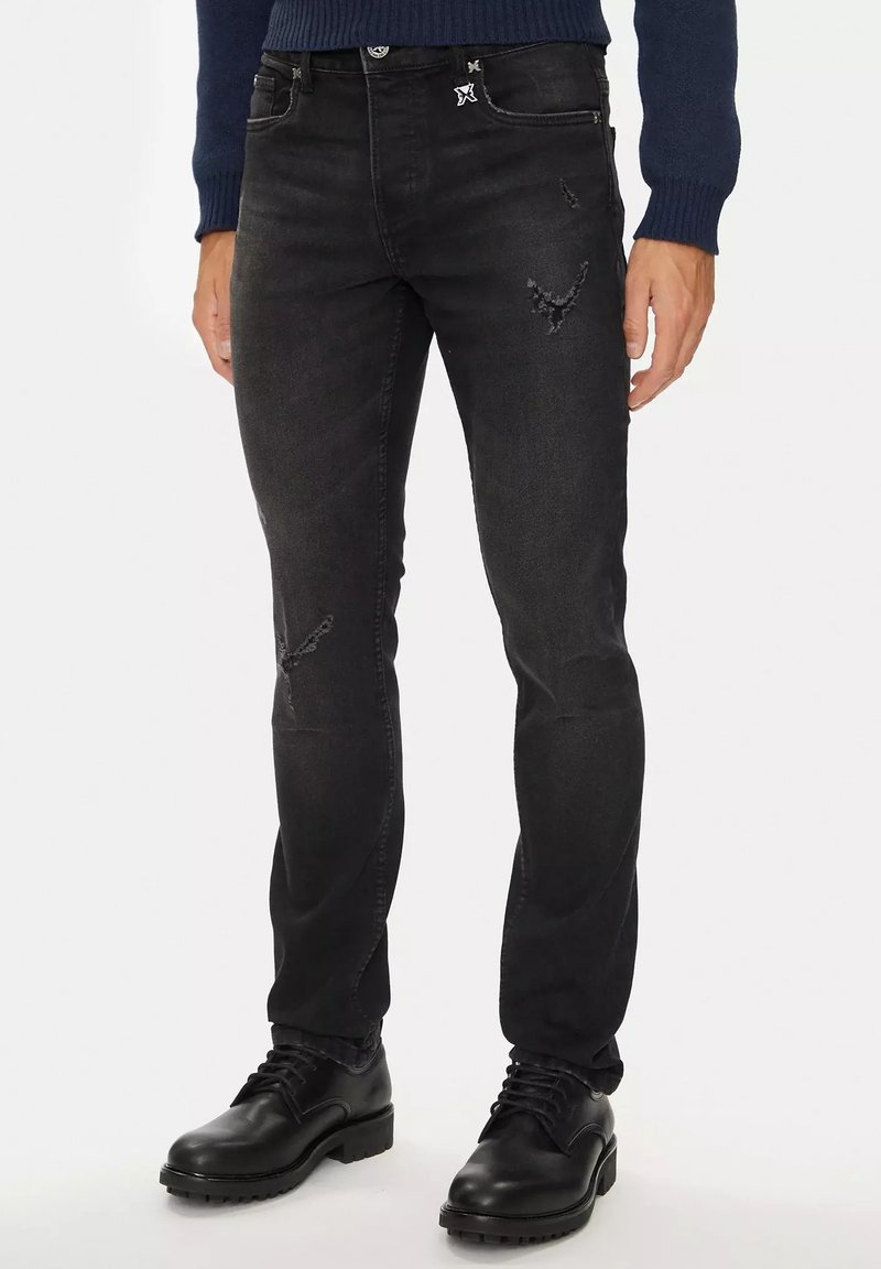 Jeans in denim nero con una vestibilità slim, caratterizzati da lievi segni di usura e un accento del logo discreto sulla tasca anteriore. Abbinati a stivali neri.