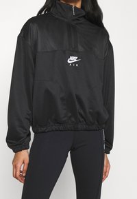Person som bär en svart Nike Air jacka med kvartdragning och elastisk nederkant samt svarta tajta leggings med vit text på sidan.
