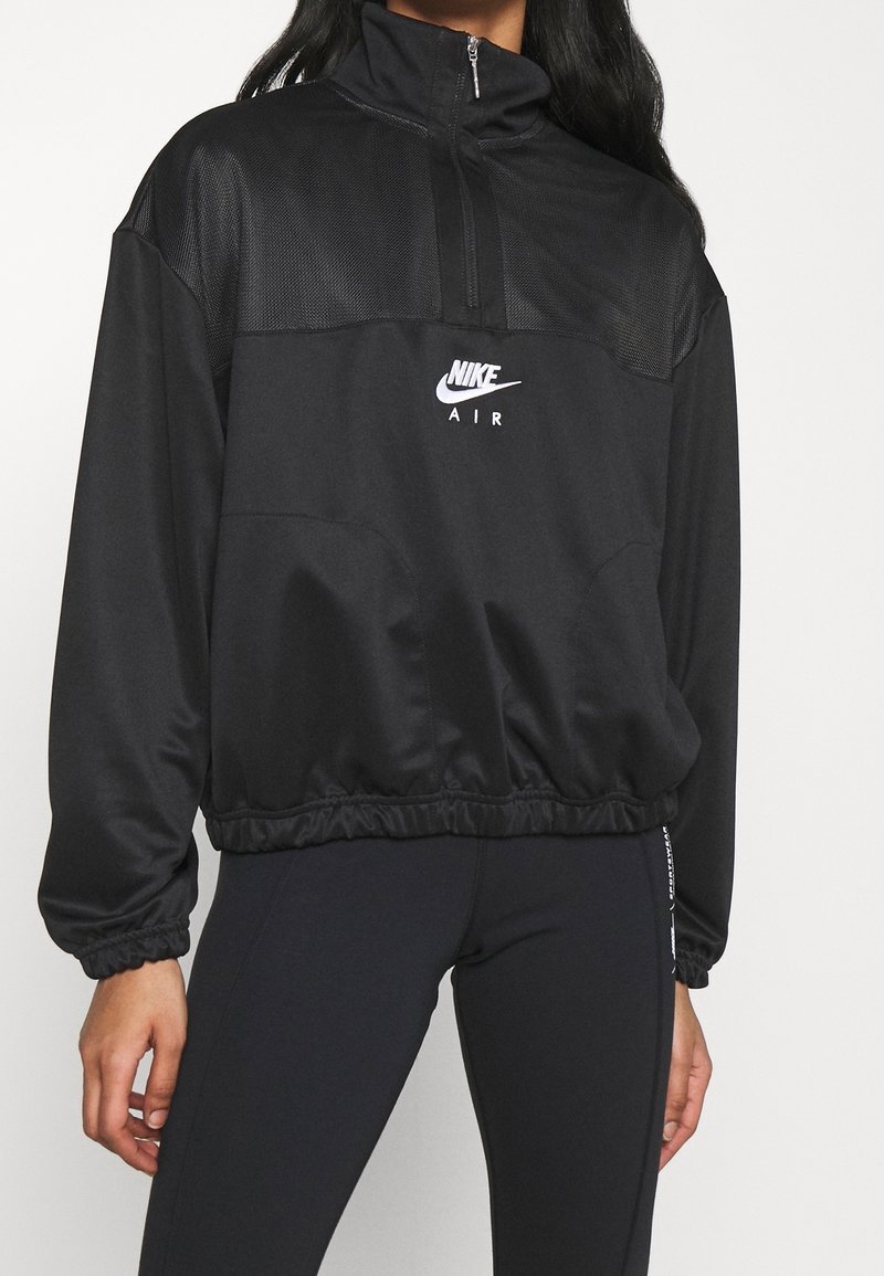 Person som bär en svart Nike Air jacka med kvartdragning och elastisk nederkant samt svarta tajta leggings med vit text på sidan.