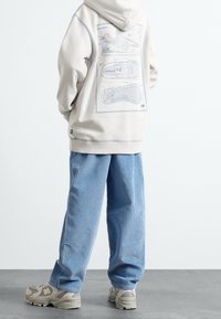 Jasnoszara bluza z kapturem z nadrukiem diagramów butów na plecach, zestawiona z niebieskimi spodniami w prawdzie. Szare trampki z teksturowaną podeszwą.