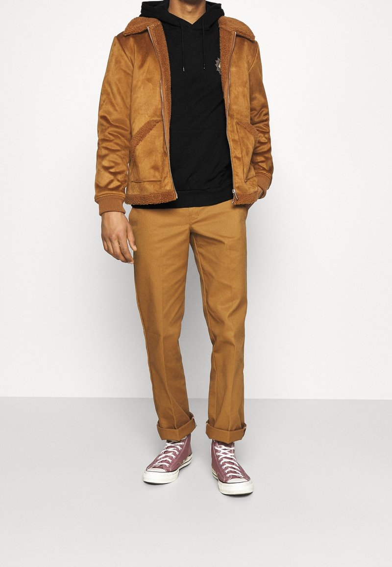 Homme portant une veste marron doublée sherpa, un sweat à capuche noir, un pantalon couleur chameau avec des revers, et des baskets montantes violettes, debout sur fond blanc.