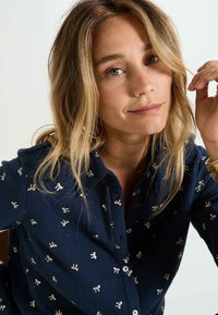 Camisa azul marino de botones con patrones de lazo dorados, tejido texturizado y cuello clásico. Modelo con cabello hasta los hombros sosteniendo un mechón.