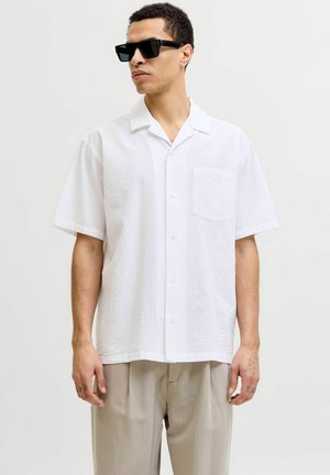 Homme portant des lunettes de soleil noires, une chemise blanche ample à manches courtes avec poche, et un pantalon beige clair à fines rayures sur fond uni.