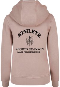 Rózsaszín kapucnis pulóver, fekete felirattal: "ATLETA", "SZAKASZ", és "Bajnokoknak készült", valamint absztrakt grafikai design.