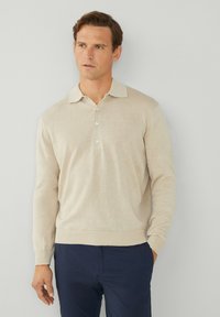 Beige Langarm-Polo-Pullover mit kragenförmigem Ausschnitt, drei-Knopf-Leiste und gerippten Abschlüssen an den Ärmeln und dem Saum. Getragen über dunkelblauen Hosen.