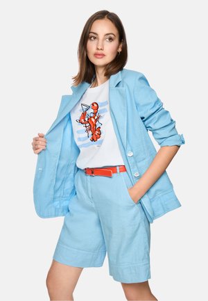 Frau trägt hellblauen Blazer und Shorts, weißes T-Shirt mit Hummer-Motiv und orangefarbenen Gürtel, posiert mit einer Hand in der Tasche.