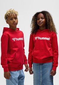 Rote Kapuzenpullover aus Baumwollmischung, mit einer Fronttasche und weißem Logo. Kombiniert mit Jeanshosen, aufgenommen in einem Studio.