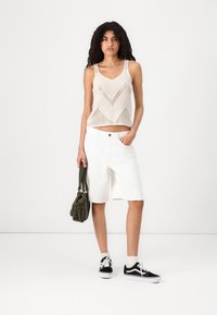 Szydełkowy top na ramiączkach w wzór zygzak w beżowym kolorze, zestawiony z białymi szortami do kolan. Modelka trzyma oliwkową torbę, nosząc czarne sneakersy.