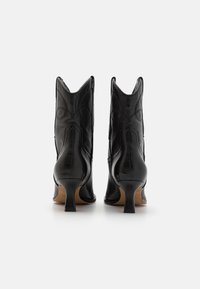 Botas de vaquero negras de cuero con tacones altos, punteras puntiagudas y costuras detalladas, que presentan un tacón delgado y un diseño distintivo con corte curvado.