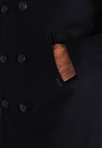 Cappotto navy doppiopetto con grandi bottoni neri, una tasca laterale e un'etichetta. Realizzato in un morbido tessuto simile alla lana.