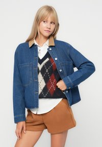Denim jacket with a brown corduroy collar, featuring two front pockets, layered over a patterned sweater and a white shirt, paired with brown shorts.