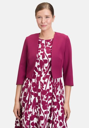 Magenta afskåret bolerojakke med trekvartærmer båret over en hvid kjole med burgunder blomsterprint og plisseret design.