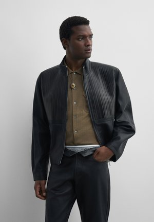 Man draagt een zwarte geplooide leren jas over een bruin overhemd met knopen, met zwarte leren broek en een hanger aan een ketting, kijkt zijwaarts.