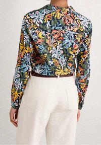Chemise à motif floral aux couleurs vives, fabriquée en tissu léger. La vue arrière montre des manches longues, un col arrondi et des fentes sur les côtés.
