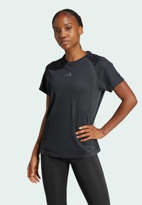 Donna con capelli intrecciati, che indossa una maglietta atletica nera e leggings, in piedi con una mano sulla vita contro uno sfondo neutro.