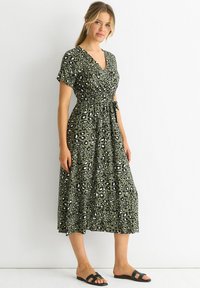 Green leopard print dress with short sleeves, cinched waist, and a tie accent. Made of lightweight fabric, falls mid-calf. Worn with black sandals.