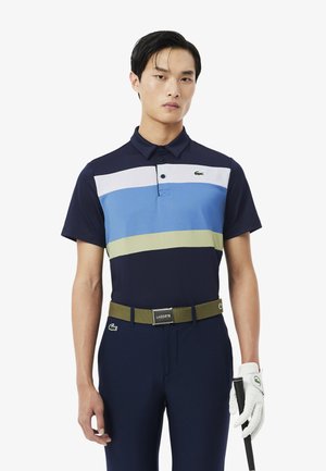 Uomo che indossa una polo blu navy con strisce bianche, blu e verdi, pantaloni blu navy, cintura oliva, guanto da golf bianco, che tiene una mazza da golf su sfondo bianco.