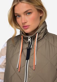 Leichte, gesteppte Weste in Taupe mit hohem Kragen. Verfügt über kontrastierende neonfarbene Kordeln, einen Frontreißverschluss und schlichte Nähte.