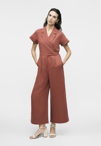 Γυναίκα στέκεται με τα χέρια στις τσέπες, φορώντας ολόσωμη φόρμα με κοντά μανίκια σε σκουριά απόχρωση και μπεζ σανδάλια με χοντρό τακούνι και λουράκια, σε λευκό φόντο.