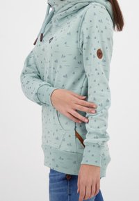 Sudadera de color azul claro hecha de algodón con pequeños patrones de veleros. Cuenta con un bolsillo canguro, puños acanalados y un parche con el logo en la manga.