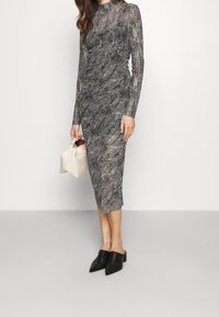 Mustavalkoinen kuviollinen pitkähiha dressi, jossa on istuva siluetti. Malli pitää käsissään vaalean kermansävyistä käsilaukkua ja antaa yhden jalan tukevana mustissa korkokenkä-muotoilussa.