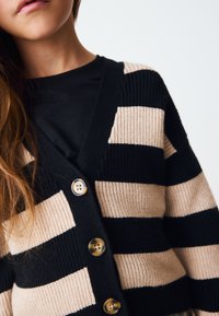 Sort og beige stribet ribstrikket cardigan med knaplukning foran og V-udskæring. Knapperne er runde og lyse i farven.