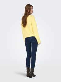 Jersey amarillo de corte holgado y mangas largas, combinado con jeans ajustados azul oscuro y botines marrones con textura.