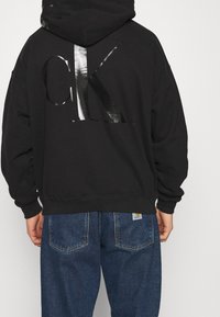 Sudadera con capucha negra y gran logotipo reflectante "CK" en la parte posterior, combinada con jeans de mezclilla azul que cuentan con bolsillos traseros y etiqueta.