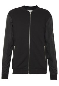 Chaqueta bomber negra con cremallera frontal. Cuerpo de algodón y mangas de nailon brillante. Cuenta con dos bolsillos laterales y un bolsillo con cremallera en la manga.