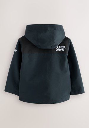 Giacca impermeabile blu navy con pannello superiore nero, cappuccio e logo "SUPERDRY" ricamato sulla schiena. Texture liscia, vestibilità standard.