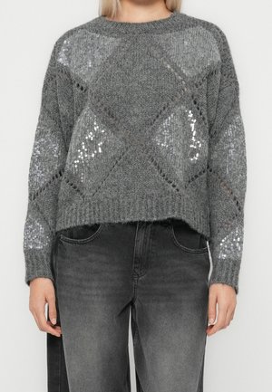 Mujer con un jersey gris de punto con patrones de lentejuelas en forma de diamante combinado con vaqueros negros desteñidos.
