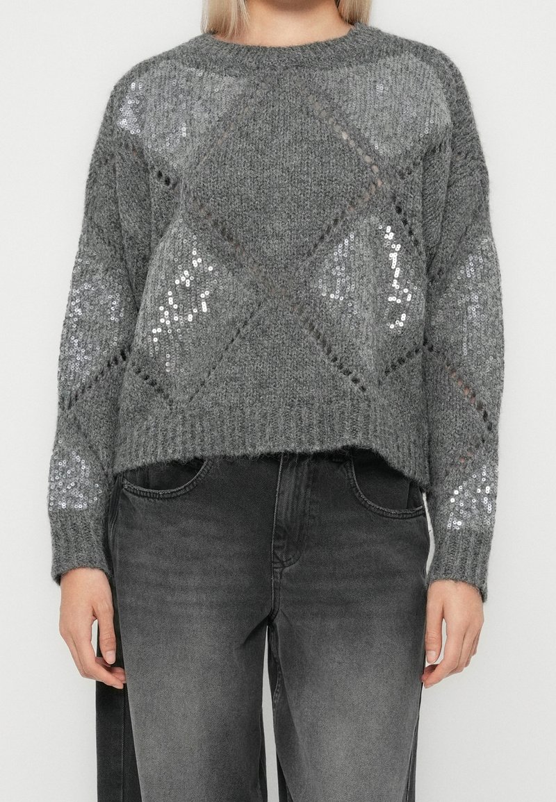Femme portant un pull en laine gris avec des motifs de sequins en forme de losange, associé à un jean noir délavé.