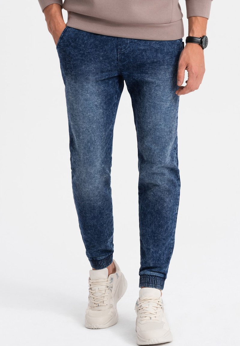 Pantalons jogger en denim bleu avec des poignets côtelés, des poches latérales et une texture usée. Assortis à des baskets de couleur claire.