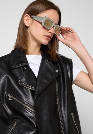 Donna con capelli castani lisci che indossa grandi occhiali da sole quadrati beige, un gilet in pelle nero e una camicia bianca a maniche corte, mentre sistema gli occhiali da sole con la mano destra.