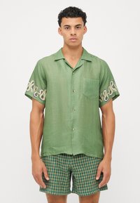 Camisa verde de manga corta con una tela texturizada, que presenta un bolsillo en el pecho y bordados en espiral en las mangas. Combinada con pantalones cortos de cuadros.