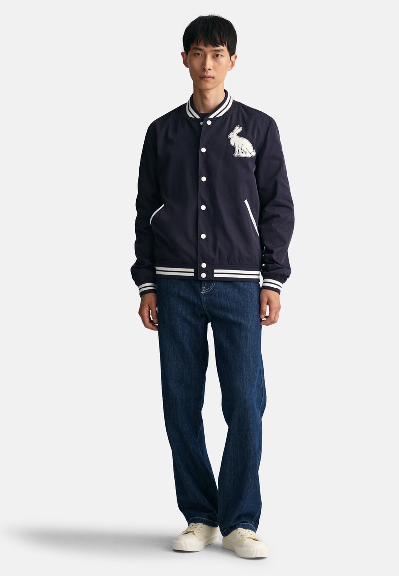 Marineblau bomberjacke mit weißen Streifenakzenten, gesticktem Hasenmotiv und Druckknöpfen, kombiniert mit geraden Jeans und hellen Sneakers.