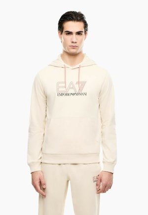Ung mand iført cremefarvet Emporio Armani hættetrøje og matchende bukser står vendt mod fronten mod en hvid baggrund.