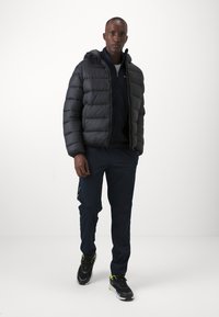 Giubbotto puffer nero con cappuccio, indossato sopra una maglia e pantaloni blu scuro. L'outfit è completato da sneakers nere e grigie con accenti neon.