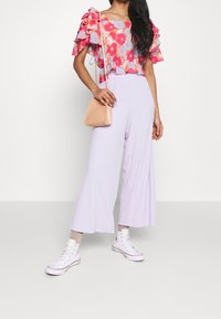 Bluse mit Blumenmuster und Rüschenärmel, kombiniert mit lavendelfarbenen, weiten Hosen und weißen Sneakers. Eine kleine, strukturierte, rosa Umhängetasche ist enthalten.