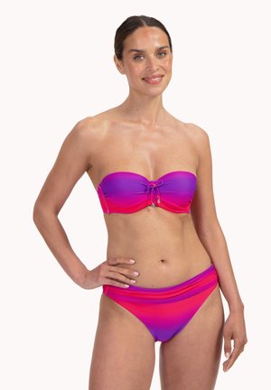 Kobieta ubrana w bikini bez ramiączek o fioletowo-różowym gradiencie z ozdobnym wiązaniem na górze, stojąca z jedną ręką na biodrze, uśmiechająca się.