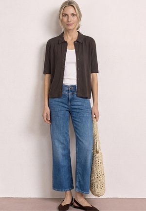 Frau steht vor einer beigen Wand, trägt ein schwarzes Kurzarmhemd mit Knöpfen, ein weißes Tanktop, blaue Jeans, braune flache Schuhe und hält eine geflochtene Tasche.