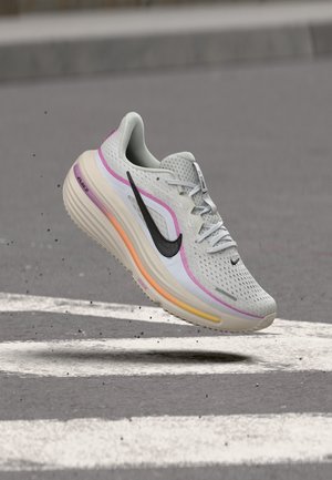 Chaussure de course blanche et rose avec le logo Nike noir flottant au-dessus d'un passage pour piétons sur une route en asphalte.