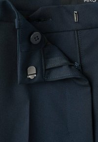 Primo piano della cintura di pantaloni blu navy che mostrano bottone, fibbia metallica, cerniera e pannello laterale elastico per una vestibilità regolabile.